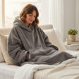 Woman wearing a gray hoodie blanket sitting on a bed in a cozy room.