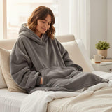 Woman wearing a gray hoodie blanket sitting on a bed in a cozy room.
