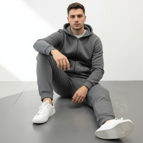 Man wearing a dark grey hoodie and pants sitting on a grey floor with a white background