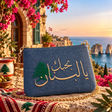 A blue textured clutch bag with gold Arabic embroidery and a green pine tree motif, resting on a patterned textile