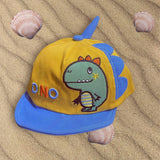 Mustard corduroy cap with blue brim, blue fabric spikes and an embroidered dinosaur, flat on beach sand