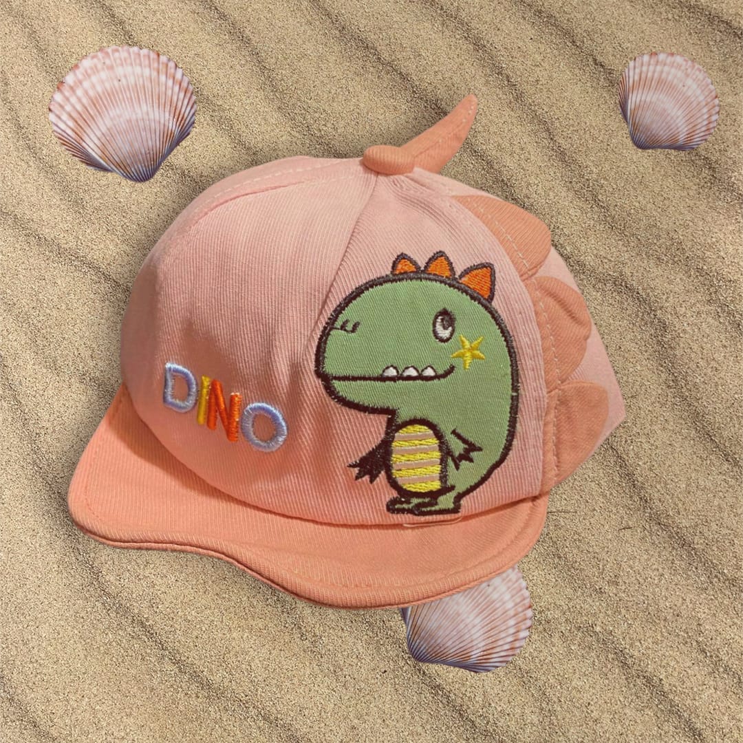 Peach corduroy kids cap with embroidered green dinosaur, side fins and a small spike, resting on sand