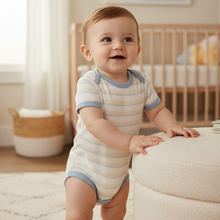 Toddler by a nursery ottoman in an off white bodysuit striped in pale blue, mustard and grey