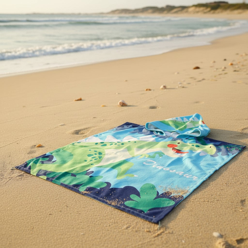 Blue dinosaur poncho towel with a green T Rex and jungle leaf print flat lay on sand