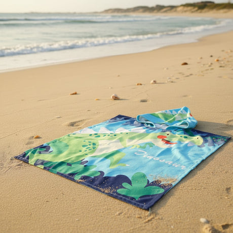 Blue dinosaur poncho towel with a green T Rex and jungle leaf print flat lay on sand
