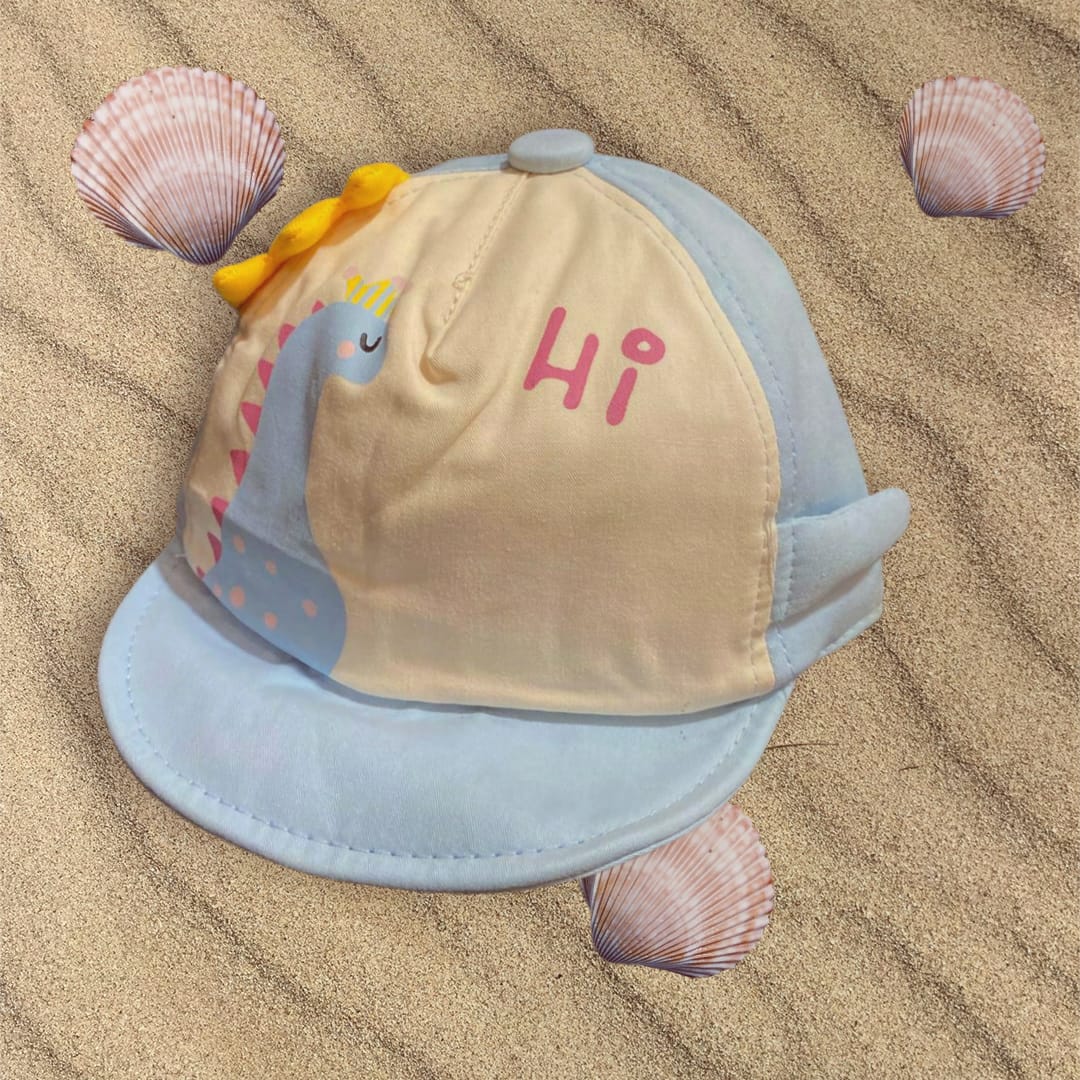 Light blue baby sun cap with cream panel, blue dinosaur print and pink Hi wording, flat on rippled sand