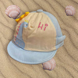 Light blue baby sun cap with cream panel, blue dinosaur print and pink Hi wording, flat on rippled sand