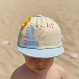 Child on sandy beach in a light blue and cream dinosaur cap with soft yellow spikes along the crown