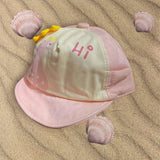 Pale pink and cream infant cap with pink dinosaur artwork and yellow crest spikes, styled beside seashells
