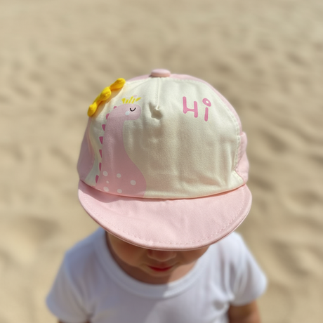 Toddler in a white tee wearing a pale pink cotton cap with a smiling dinosaur print and short brim