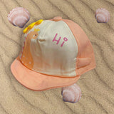 Salmon and cream kids sun cap with a printed dinosaur, yellow spikes and pink Hi text, laid flat on sand