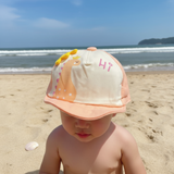 Toddler at the beach wearing a salmon and cream baby cap with a printed dinosaur and pink Hi lettering