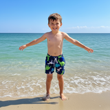Child in colorful dinosaur design swim trunks standing on a beach with ocean in the background