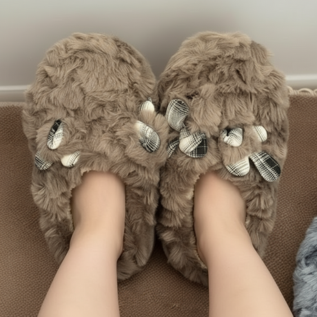 Taupe fluffy slippers with checked fabric bows worn on feet resting on a brown sofa
