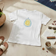 A white short-sleeve T-shirt laid flat on a light wood floor, featuring a colorful avocado graphic on the chest. The shirt is styled in a cozy room setting with a potted plant, slide sandals, sunglasses on an open book, a cup of tea, and a patterned rug nearby.