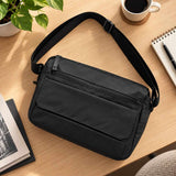 Black crossbody messenger bag with a flap pocket and long zip laid on a desk beside a notebook