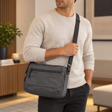 Man in a cream sweater wearing the grey crossbody bag on one shoulder in a warm living room
