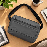 Grey crossbody messenger bag with a front flap pocket and black strap on a wooden desk with coffee