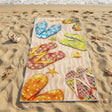 A rectangular beach towel laid flat on sandy shore near the ocean, printed with colorful flip-flop sandals and small starfish motifs. The towel is surrounded by sand, shells, and seaweed, with waves visible in the background.