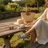 Woman sitting at a wooden table outdoors with a straw bag, hat, and floral handmade sandals