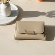 Beige wallet with gold clasp on a light wooden surface, with a plant vase in the background