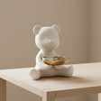 White ceramic bear figurine with a gold dish on a wooden table against a beige wall.