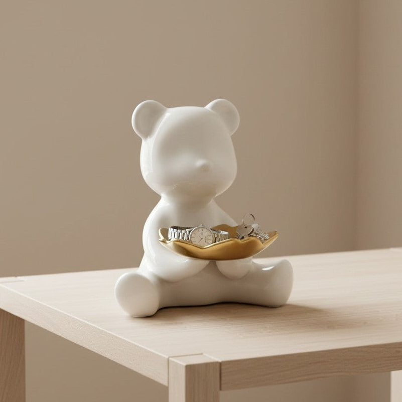 White ceramic bear figurine with a gold dish on a wooden table against a beige wall.