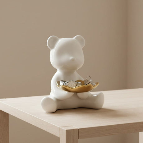 White ceramic bear figurine with a gold dish on a wooden table against a beige wall.