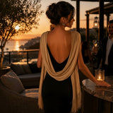 A woman stands on a rooftop terrace at sunset wearing a black backless dress with a draped gold shawl over her shoulders