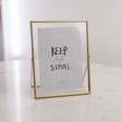 Close view of one slim gold floating frame on white marble, its easel leg behind