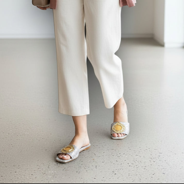 Person wearing white pants, and gold ring handmade sandals in a minimal indoor setting