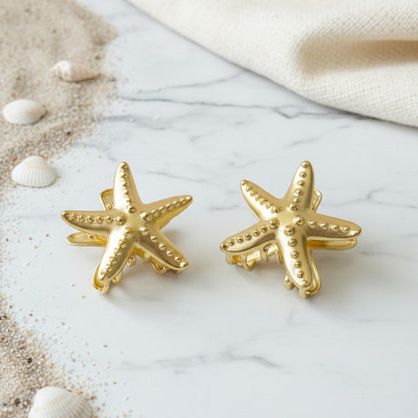 Two polished gold starfish claw clips on marble with small seashells