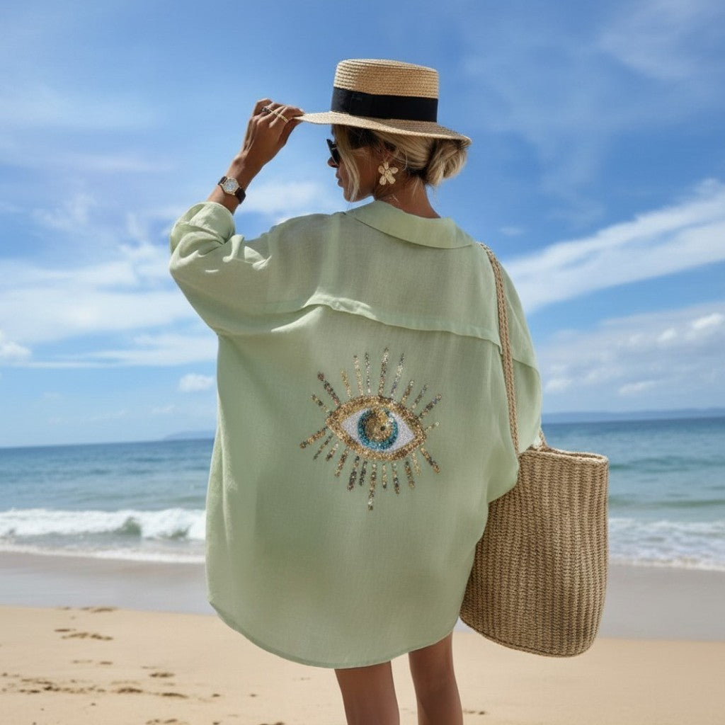 Person on a beach wearing a light green shirt with an embroidered design, holding a straw hat, and carrying a woven bag.