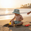 A toddler sitting on a sandy beach at sunset, wearing a green camouflage bucket hat and matching swim trunks