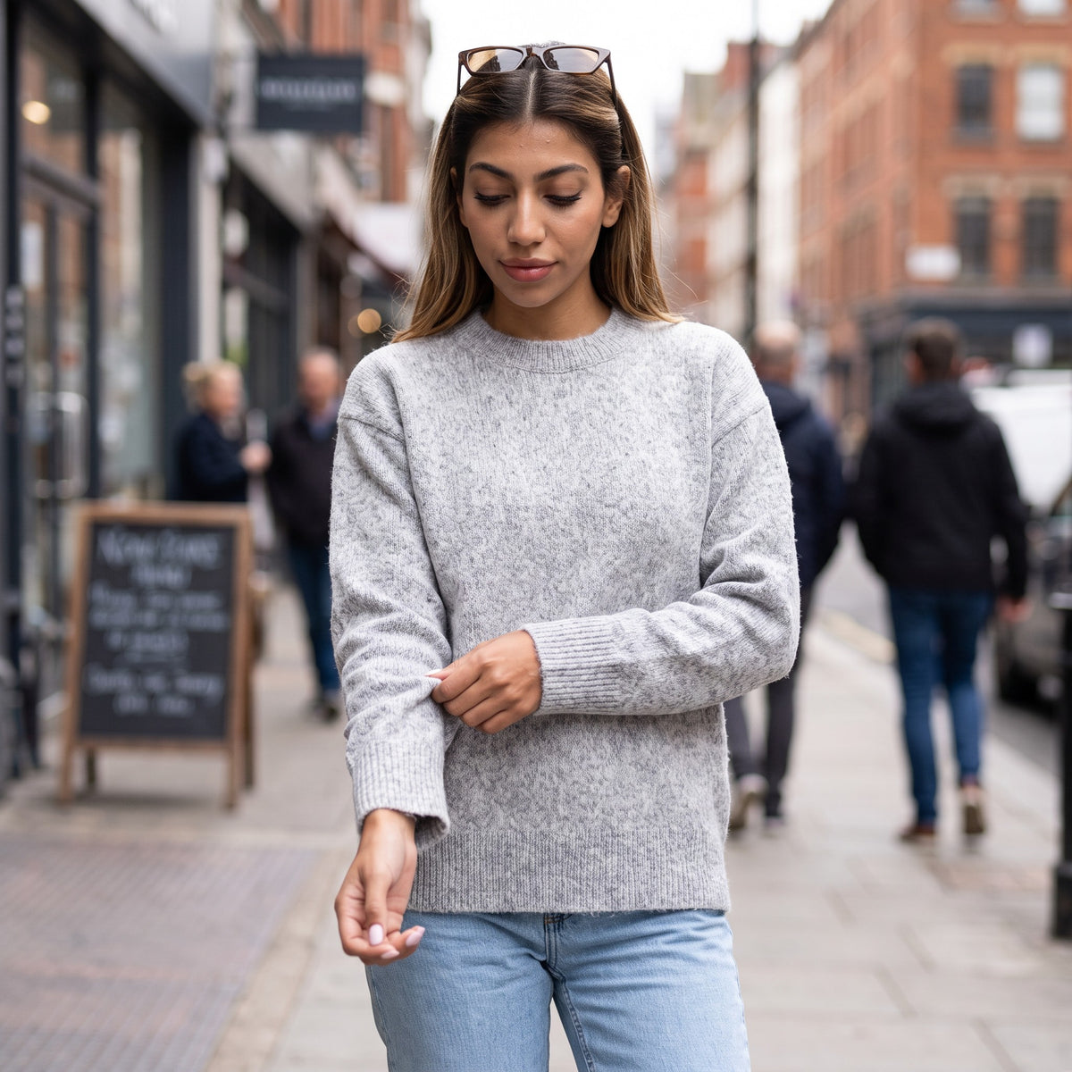 Woman in a grey brushed wool crew neck sweater and light jeans adjusting her sleeve on a busy shopping street
