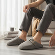 Man wearing grey corduroy slippers with a gold toned buckle strap across the vamp