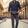 Man walking down a sunlit street wearing a blue shirt, grey pants, and a waist bag