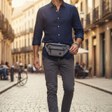 Man walking down a sunlit street wearing a blue shirt, grey pants, and a waist bag
