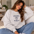 Woman in a grey sweatshirt with a vintage heart crest print on the chest, relaxing with jeans