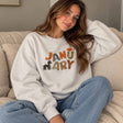 Woman in a grey crew neck sweatshirt with colourful January lettering and a puppy print, sitting in jeans