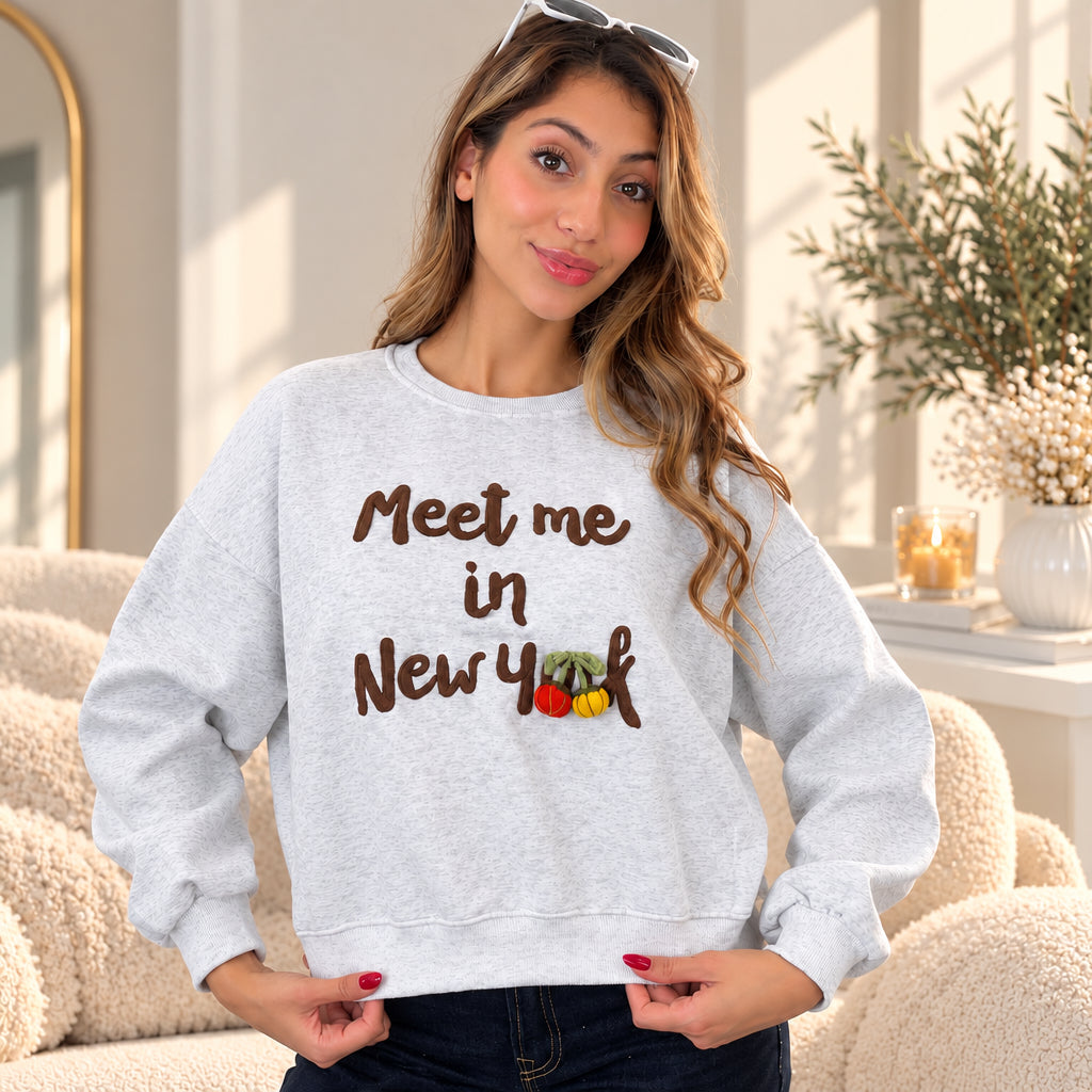 Woman with sunglasses on her head in a heather grey Meet me in New York sweatshirt with brown lettering