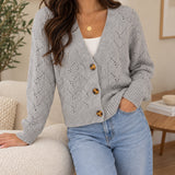 Grey pointelle knit cardigan with three tortoiseshell buttons, worn leaning on a boucle sofa