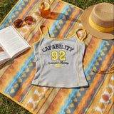 Tank top with 'CAPABILITY 92' text on a colorful blanket with a book, sunglasses, and hat.