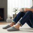 Man wearing grey waffle knit slippers on a soft rug while holding a mug