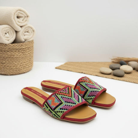 Colorful handmade slippers with geometric pattern on a white surface with a basket of towels and stones in the background