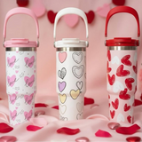 Three heart print travel mugs with flip lids and carry handles on a pink backdrop