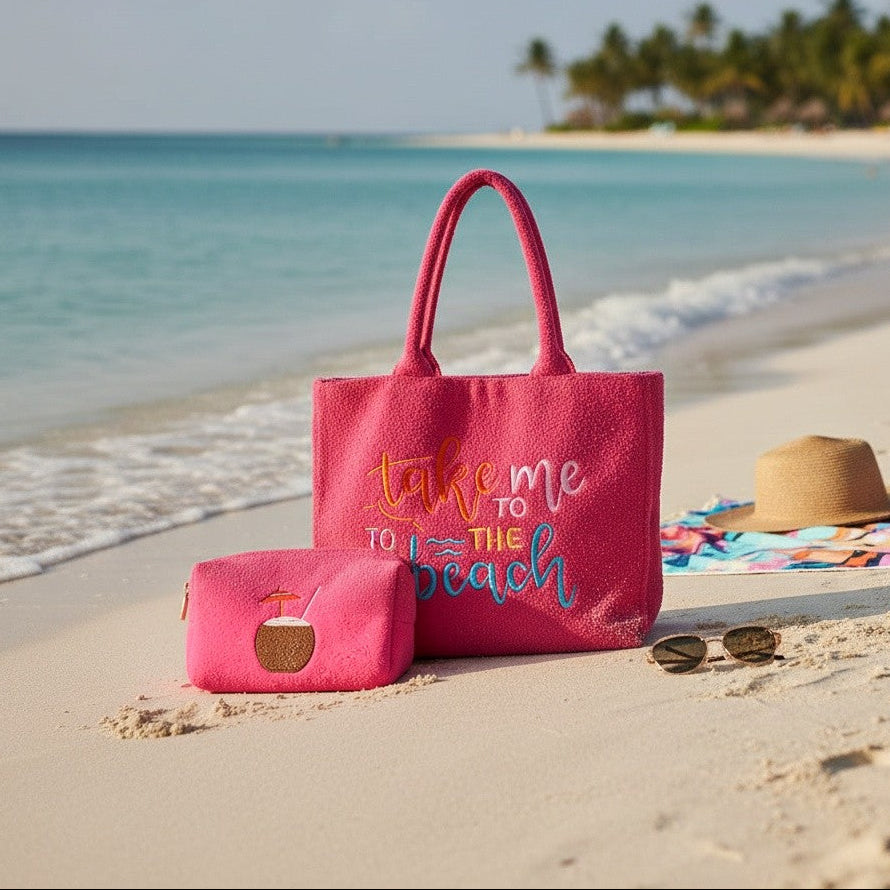 Pink tote bag with 'take me to the beach' text on a sandy beach with ocean and palm trees in the background.