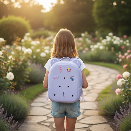 Girl on a garden path with a pale lavender hard shell backpack holding flamingo, rainbow and flower charms