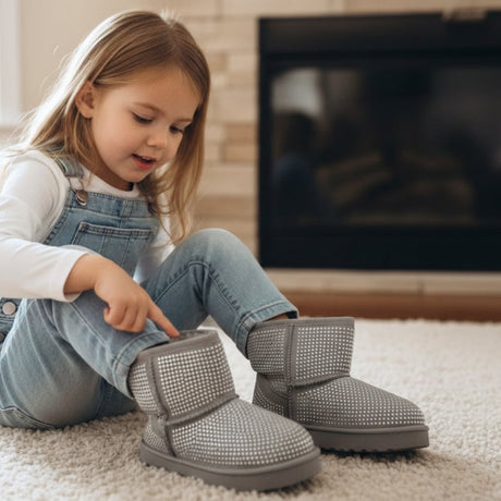 Girl pulling on grey kids winter boots by a fireplace