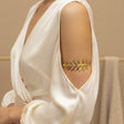 Gold leaf bracelet on a person wearing a white dress against a beige background